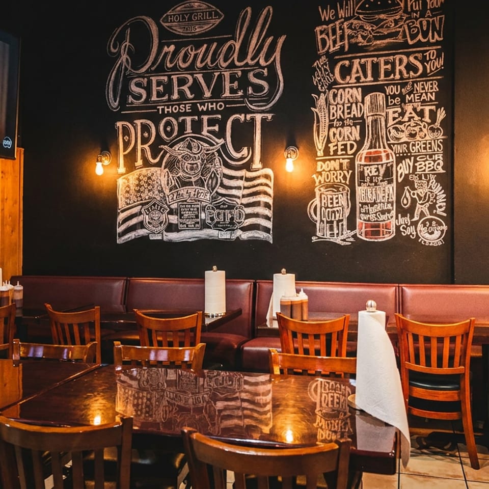 Restaurant interior with wooden tables, booths, and a chalkboard wall with "Holy Grill" branding.