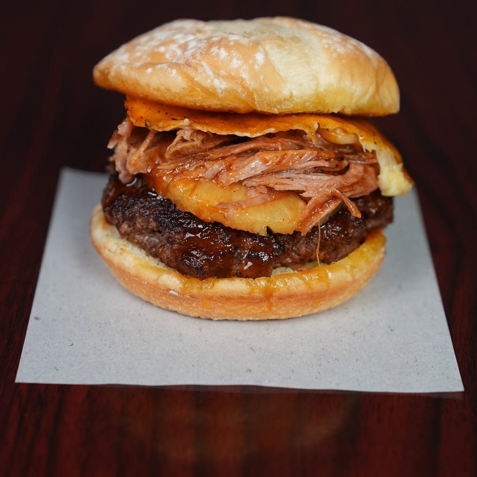 Close-up of a loaded burger with a beef patty, pulled meat, fried egg, and toasted bun on a plate.