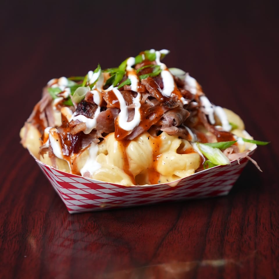 Loaded fries topped with melted cheese, sliced meat, green onions, herbs, and white and orange sauces in a paper tray.